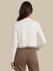 A woman shown from the back wearing a cream ribbed knit cropped cardigan with a sheer ruffle hem, paired with brown pants, against a solid beige background.