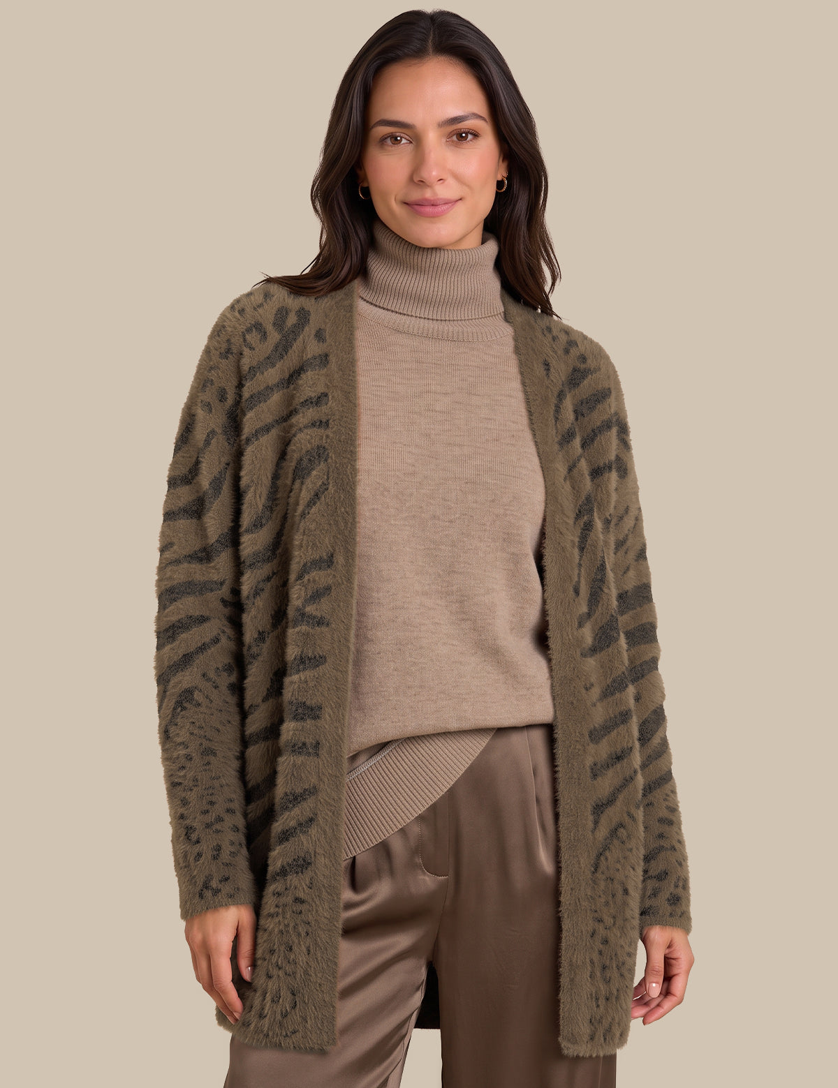 A woman modeling a fuzzy open-front cardigan with a dark gray zebra-like pattern on an olive green base, worn over a beige turtleneck sweater and brown pants, against a solid beige background.