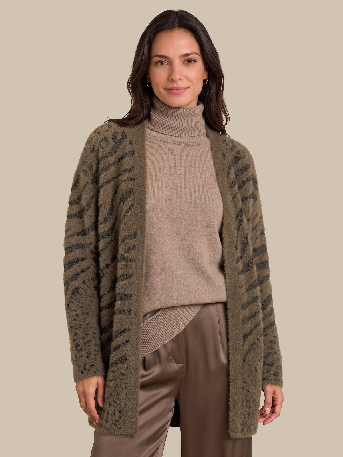A woman modeling a fuzzy open-front cardigan with a dark gray zebra-like pattern on an olive green base, worn over a beige turtleneck sweater and brown pants, against a solid beige background.