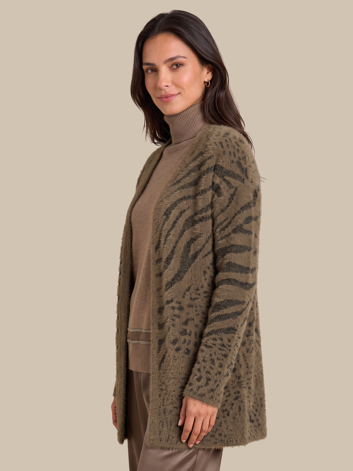 A woman modeling a fuzzy open-front cardigan with an olive-brown and dark gray animal-stripe pattern, worn over a taupe turtleneck sweater and matching pants, set against a solid beige background.