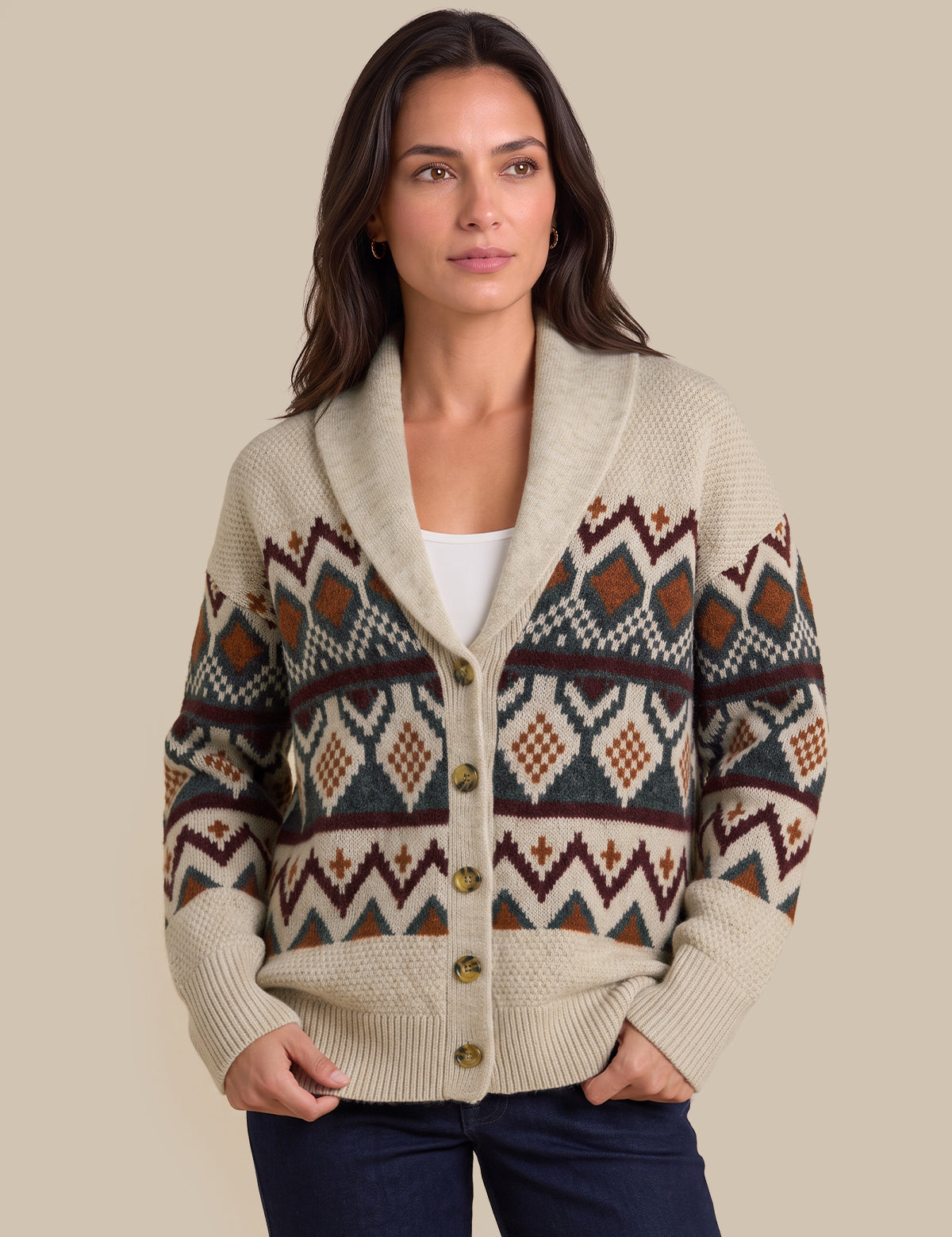 A woman wearing a cream-colored knit cardigan with a colorful geometric Fair Isle pattern in brown, rust, and navy, styled with a white top and dark blue jeans against a solid beige background.