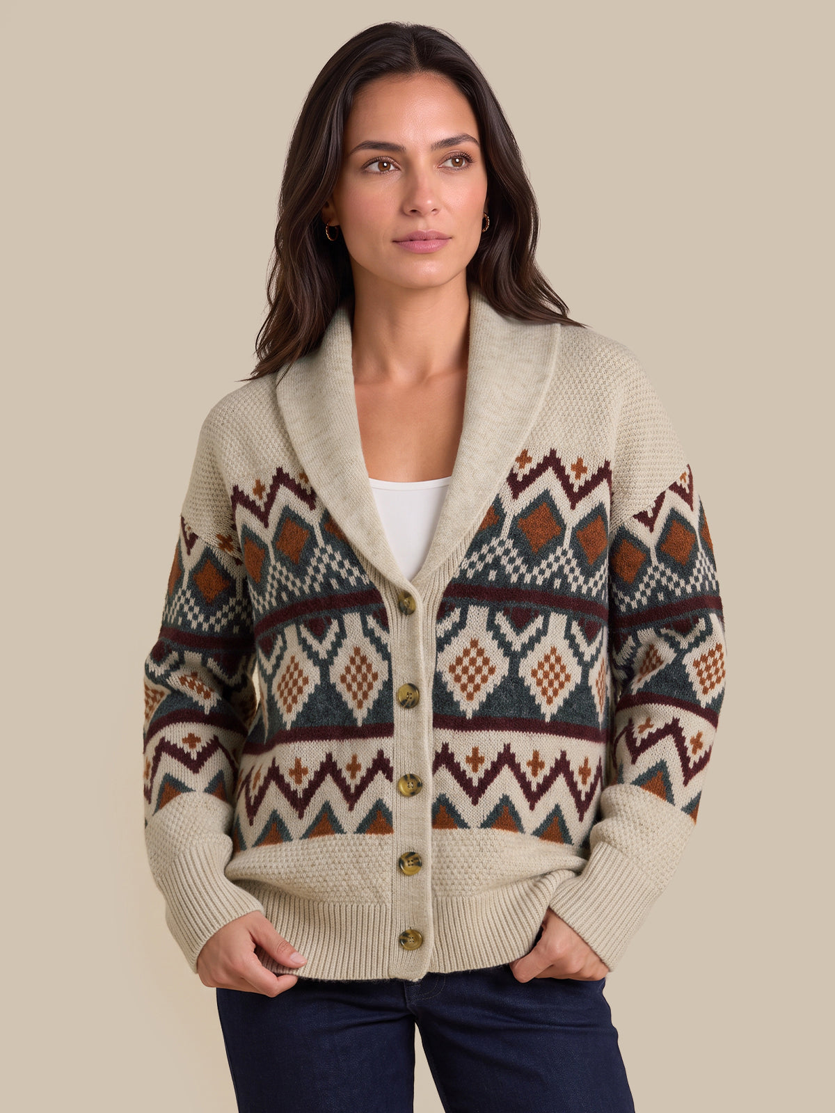 A woman wearing a cream-colored knit cardigan with a colorful geometric Fair Isle pattern in brown, rust, and navy, styled with a white top and dark blue jeans against a solid beige background.
