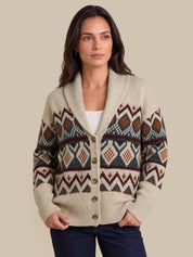 A woman wearing a cream-colored knit cardigan with a colorful geometric Fair Isle pattern in brown, rust, and navy, styled with a white top and dark blue jeans against a solid beige background.