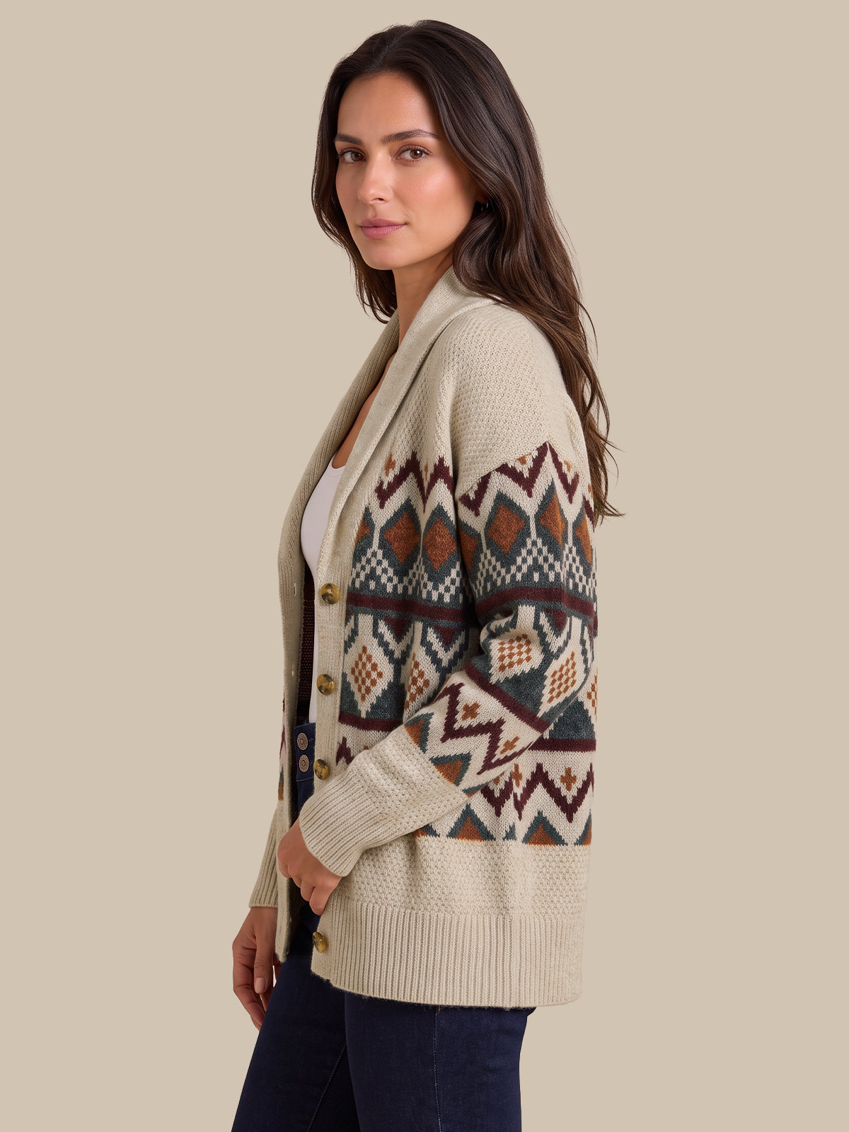 A woman wearing a cream-colored knit cardigan with a colorful geometric pattern on the sleeves and chest, paired with dark blue jeans, set against a solid beige background.