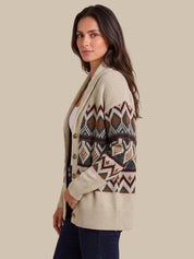 A woman wearing a cream-colored knit cardigan with a colorful geometric pattern on the sleeves and chest, paired with dark blue jeans, set against a solid beige background.
