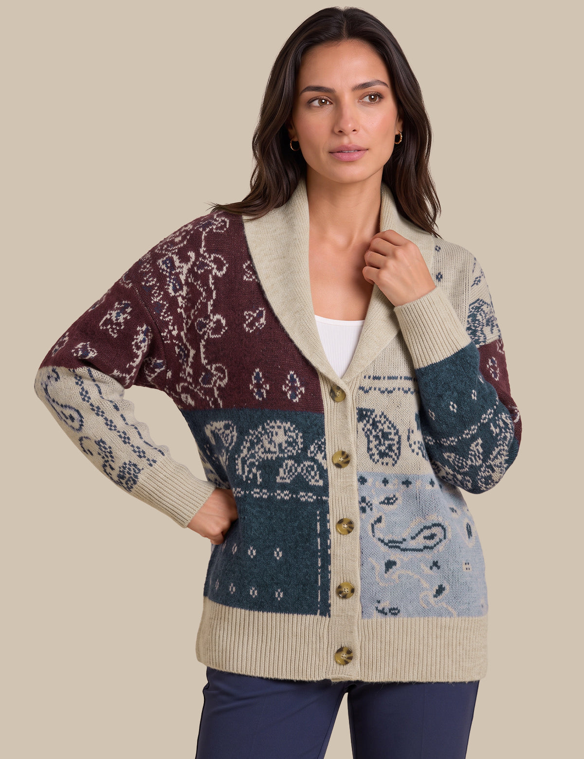 A woman modeling a multicolor patchwork knit cardigan with cream, burgundy, teal, and light blue sections, featuring a shawl collar and button-front closure, against a solid beige background.