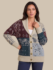 A woman modeling a multicolor patchwork knit cardigan with cream, burgundy, teal, and light blue sections, featuring a shawl collar and button-front closure, against a solid beige background.