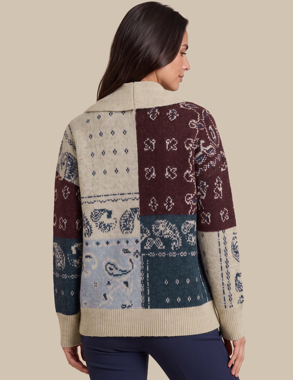A woman shown from the back wearing a patchwork knit sweater with bandana and paisley patterns in beige, burgundy, blue, and teal, paired with navy pants, against a solid beige background.