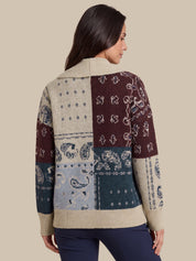 A woman shown from the back wearing a patchwork knit sweater with bandana and paisley patterns in beige, burgundy, blue, and teal, paired with navy pants, against a solid beige background.
