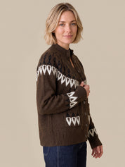 A woman wearing a brown button-front knit cardigan with white geometric patterns, paired with dark blue jeans, set against a solid beige background.