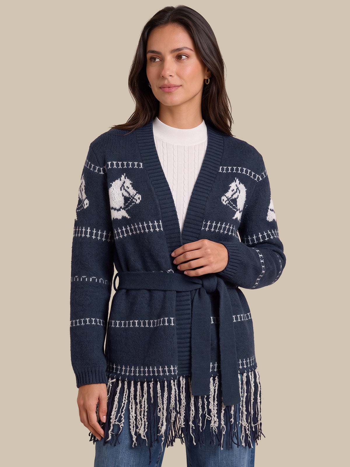 A woman wearing a navy blue knit cardigan with white horse motifs and fringe trim, styled over a white top and blue jeans, against a solid beige background.