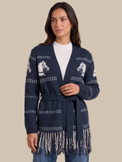 A woman wearing a navy blue knit cardigan with white horse motifs and fringe trim, styled over a white top and blue jeans, against a solid beige background.