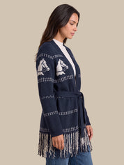 A woman wearing a navy blue knit cardigan with white horse motifs and fringe trim, shown in a side profile against a solid beige background.