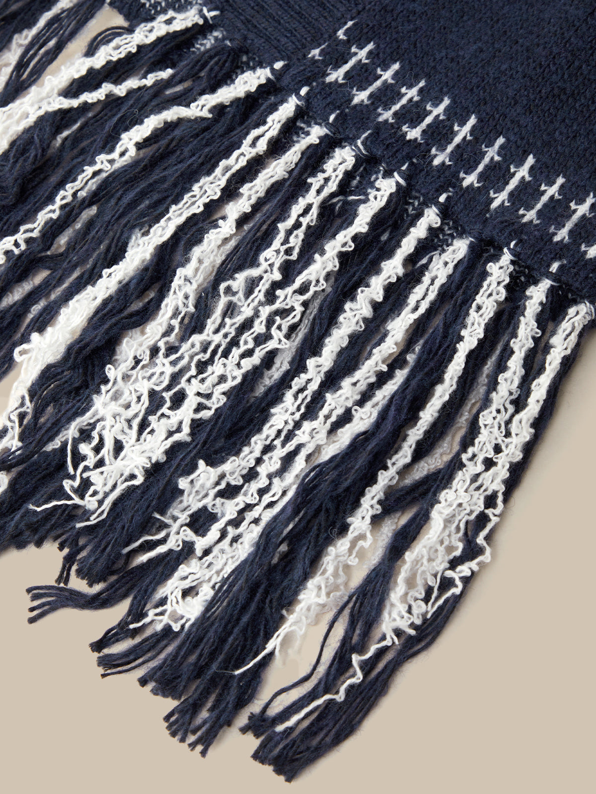 Close-up of a woven textile with dark navy blue and off-white stripes and long fringe tassels, shown against a plain beige background.