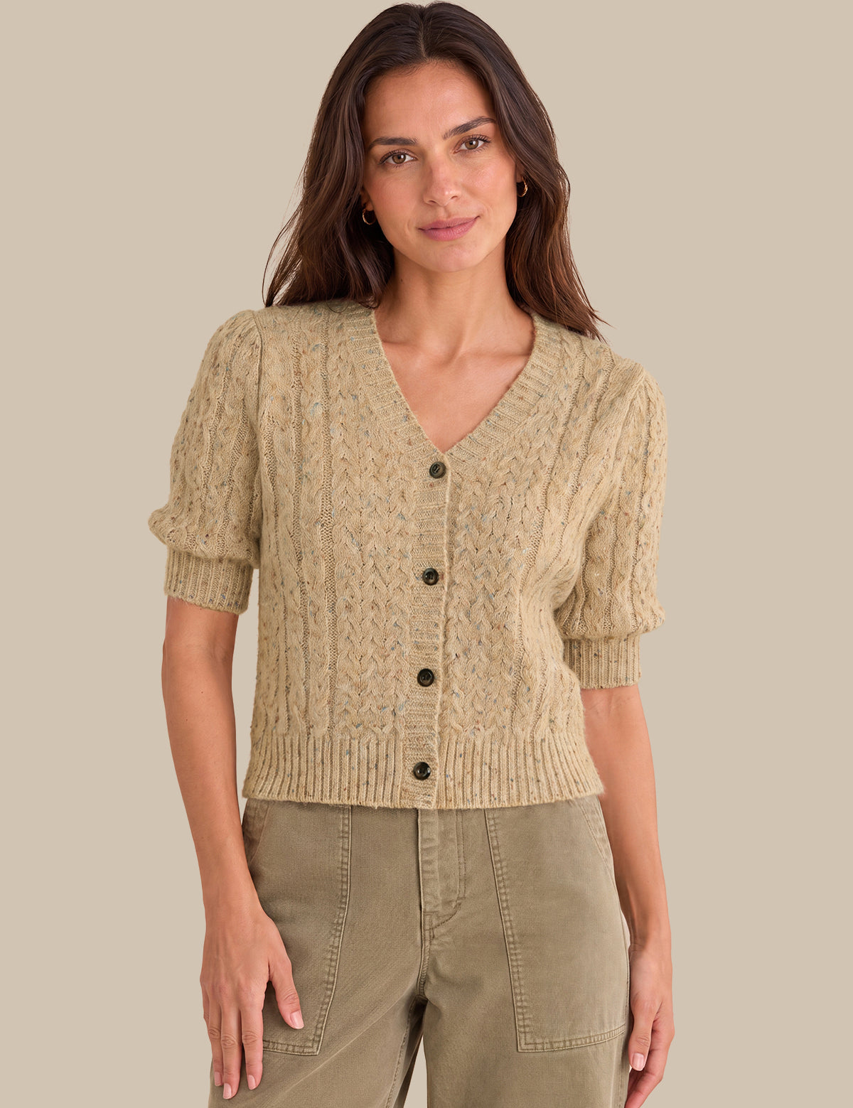 A woman modeling a short-sleeve cable-knit cardigan in a light beige color, paired with olive green pants, against a solid beige background.