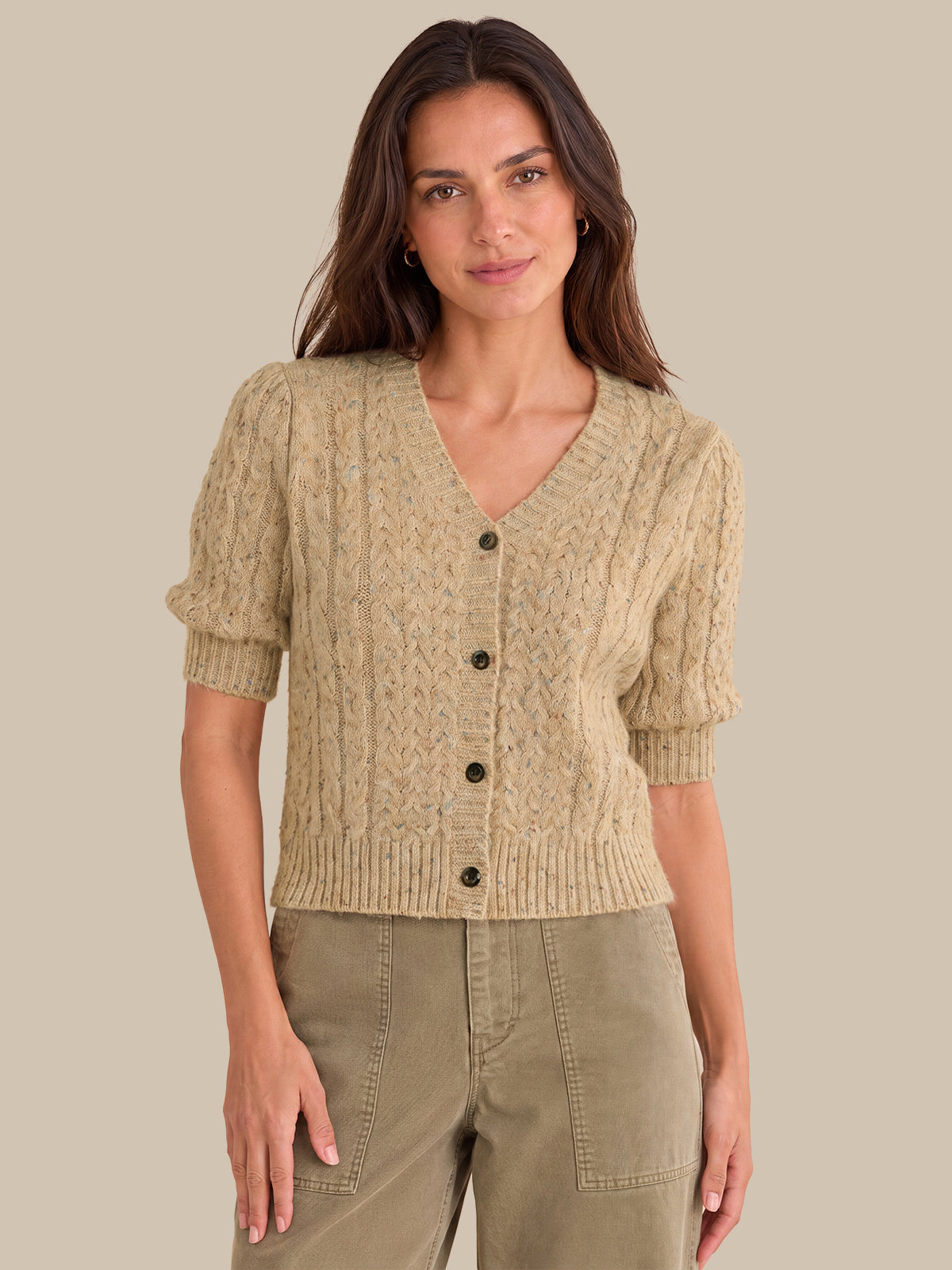 A woman modeling a short-sleeve cable-knit cardigan in a light beige color, paired with olive green pants, against a solid beige background.