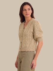 A woman modeling a cropped, short puff-sleeve cardigan in a textured beige cable knit, paired with khaki pants, against a solid beige background.