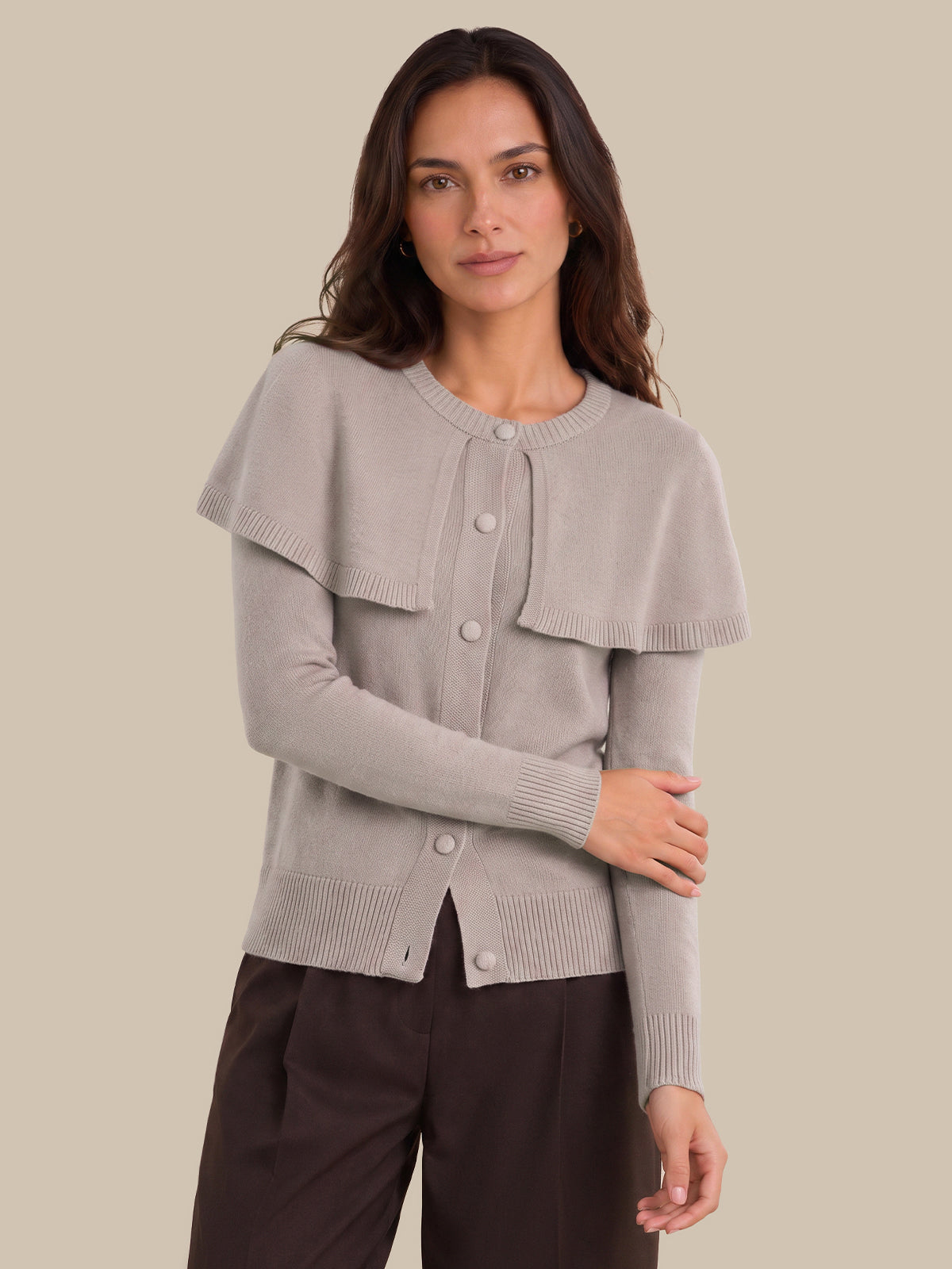 A woman modeling a light taupe knit cardigan with a unique layered ruffle collar and front buttons, paired with dark brown pants, against a solid beige background.