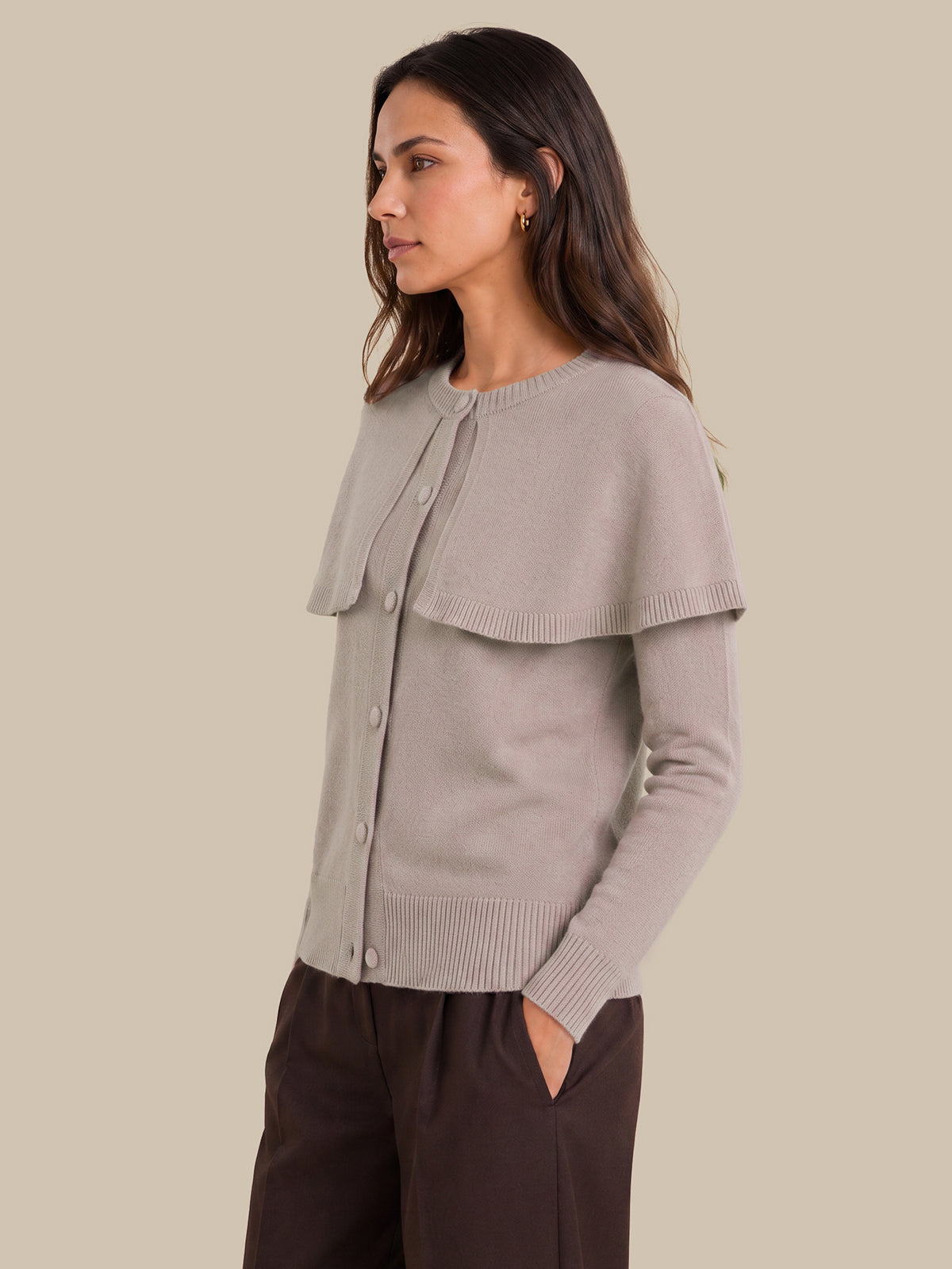 A woman wearing a light taupe knit cardigan with a wide draped yoke and dark brown trousers, shown in a side profile against a solid beige background.