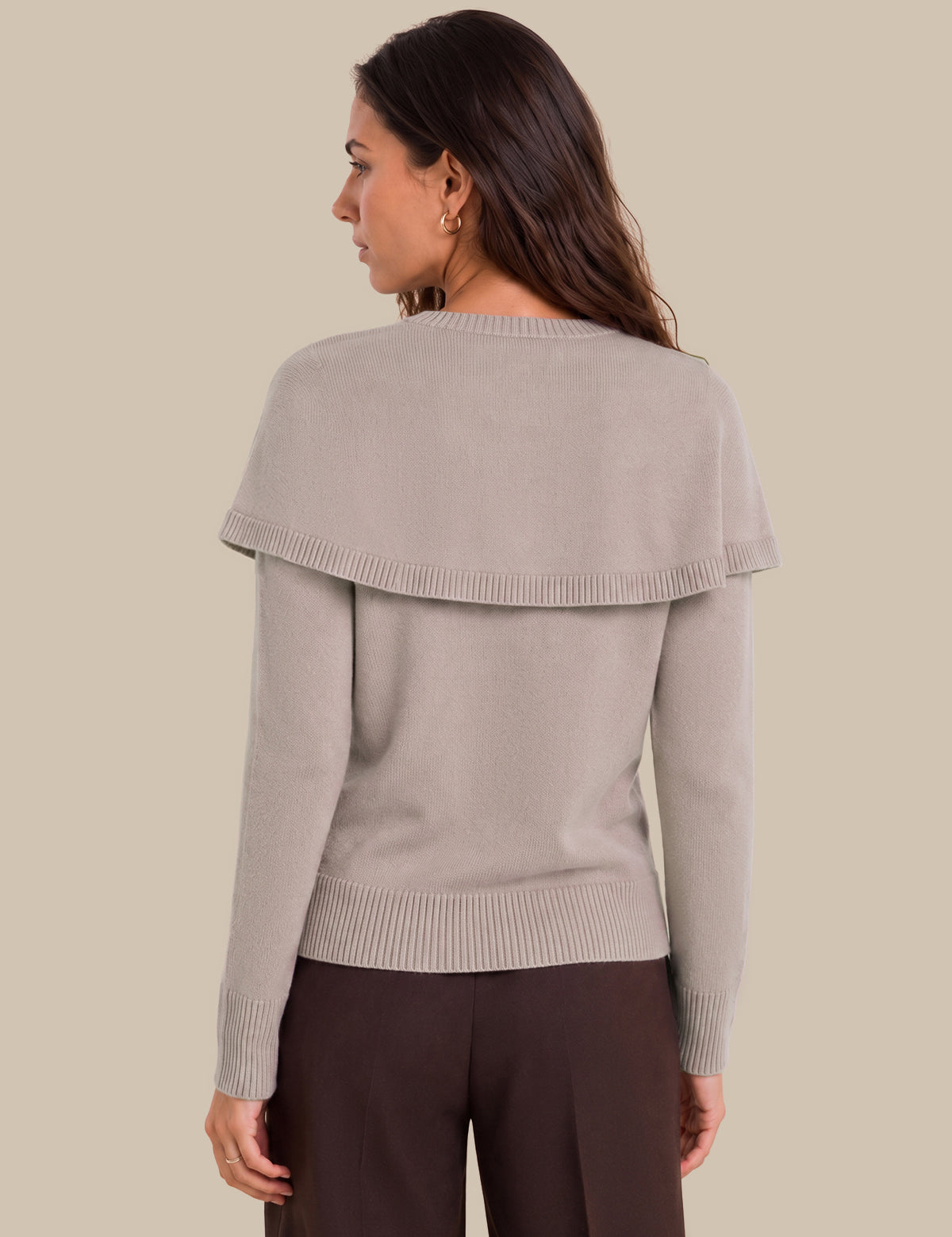 A woman shown from the back wearing a light taupe knit sweater with a wide draped shoulder detail and dark brown trousers, set against a solid beige background.
