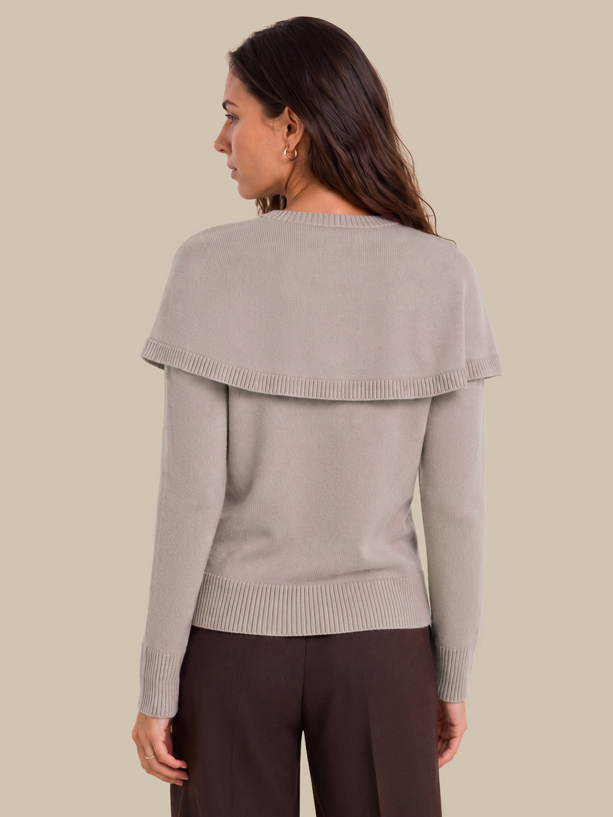 A woman shown from the back wearing a light taupe knit sweater with a wide draped shoulder detail and dark brown trousers, set against a solid beige background.