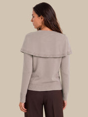 A woman shown from the back wearing a light taupe knit sweater with a wide draped shoulder detail and dark brown trousers, set against a solid beige background.