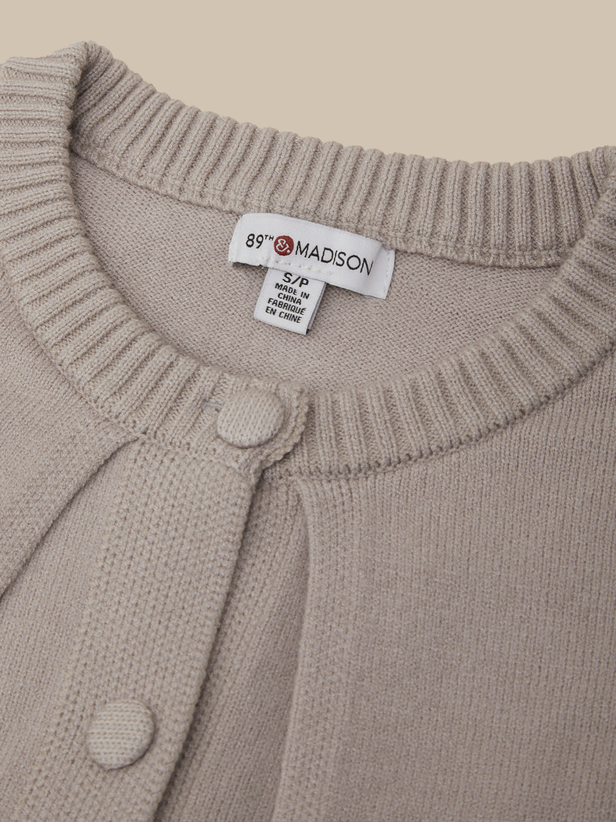 Close-up of a beige knit cardigan sweater showing the ribbed collar, a white interior neck label that reads 89 MADSON, and two matching fabric buttons.