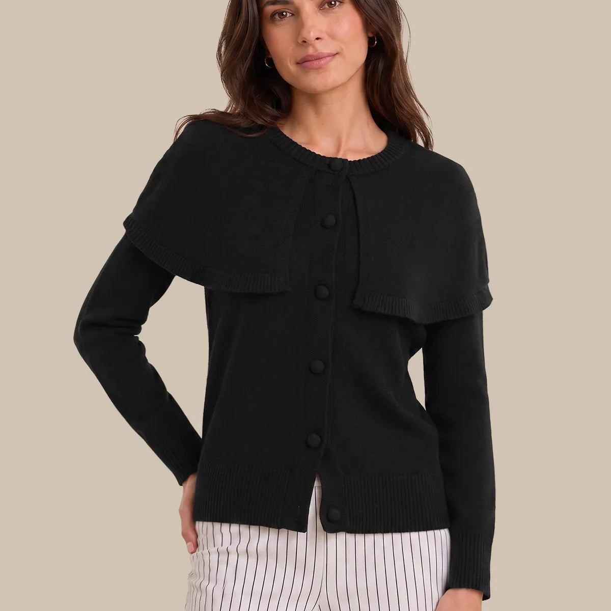 A woman modeling a black long-sleeve knit cardigan with a unique draped collar and front button closure, paired with white pinstriped pants, against a solid beige background.