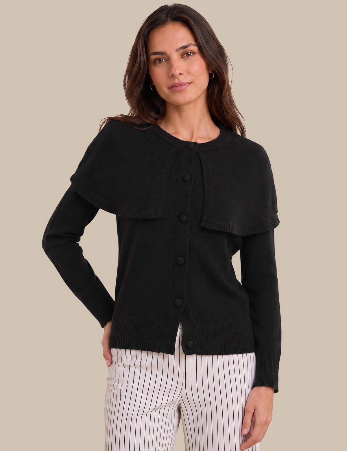 A woman modeling a black long-sleeve knit cardigan with a unique draped collar and front button closure, paired with white pinstriped pants, against a solid beige background.