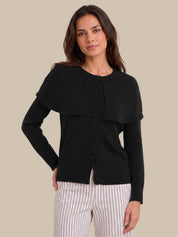 A woman modeling a black long-sleeve knit cardigan with a unique draped collar and front button closure, paired with white pinstriped pants, against a solid beige background.