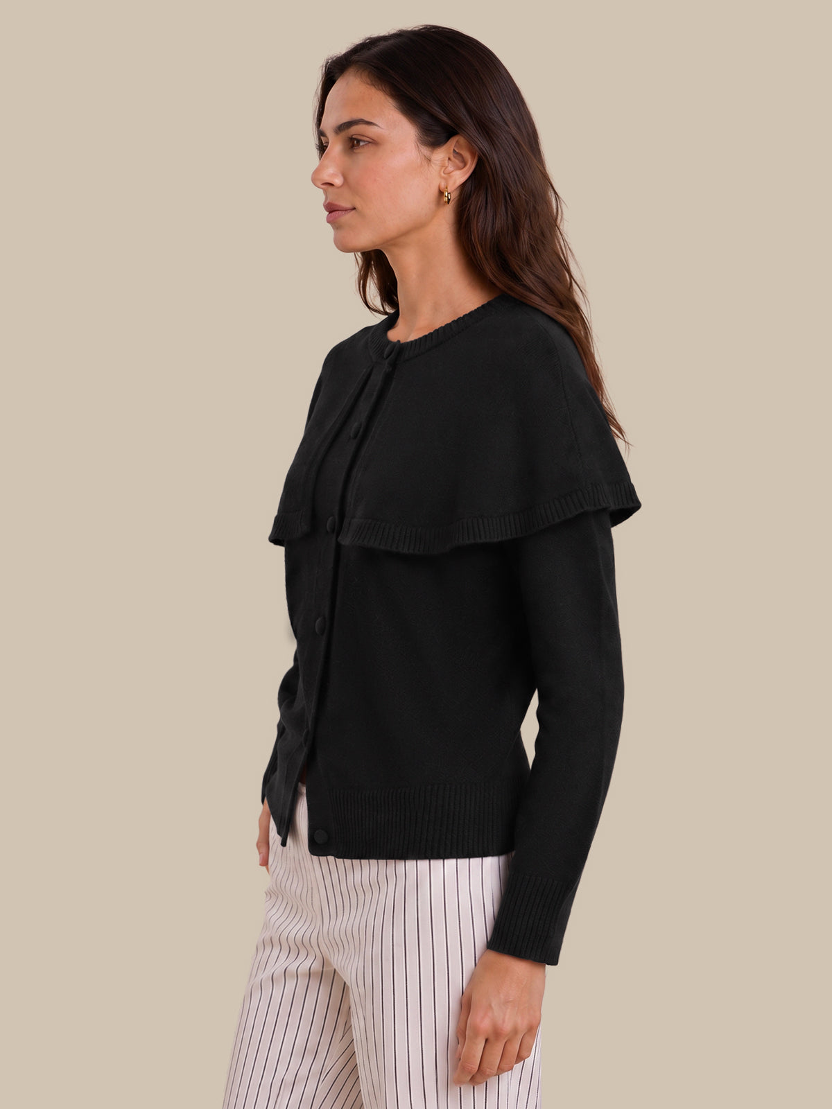 A woman shown in side profile wearing a black long-sleeve knit cardigan with a wide layered collar and white pinstriped trousers, against a solid beige studio background.