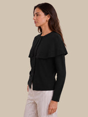 A woman shown in side profile wearing a black long-sleeve knit cardigan with a wide layered collar and white pinstriped trousers, against a solid beige studio background.
