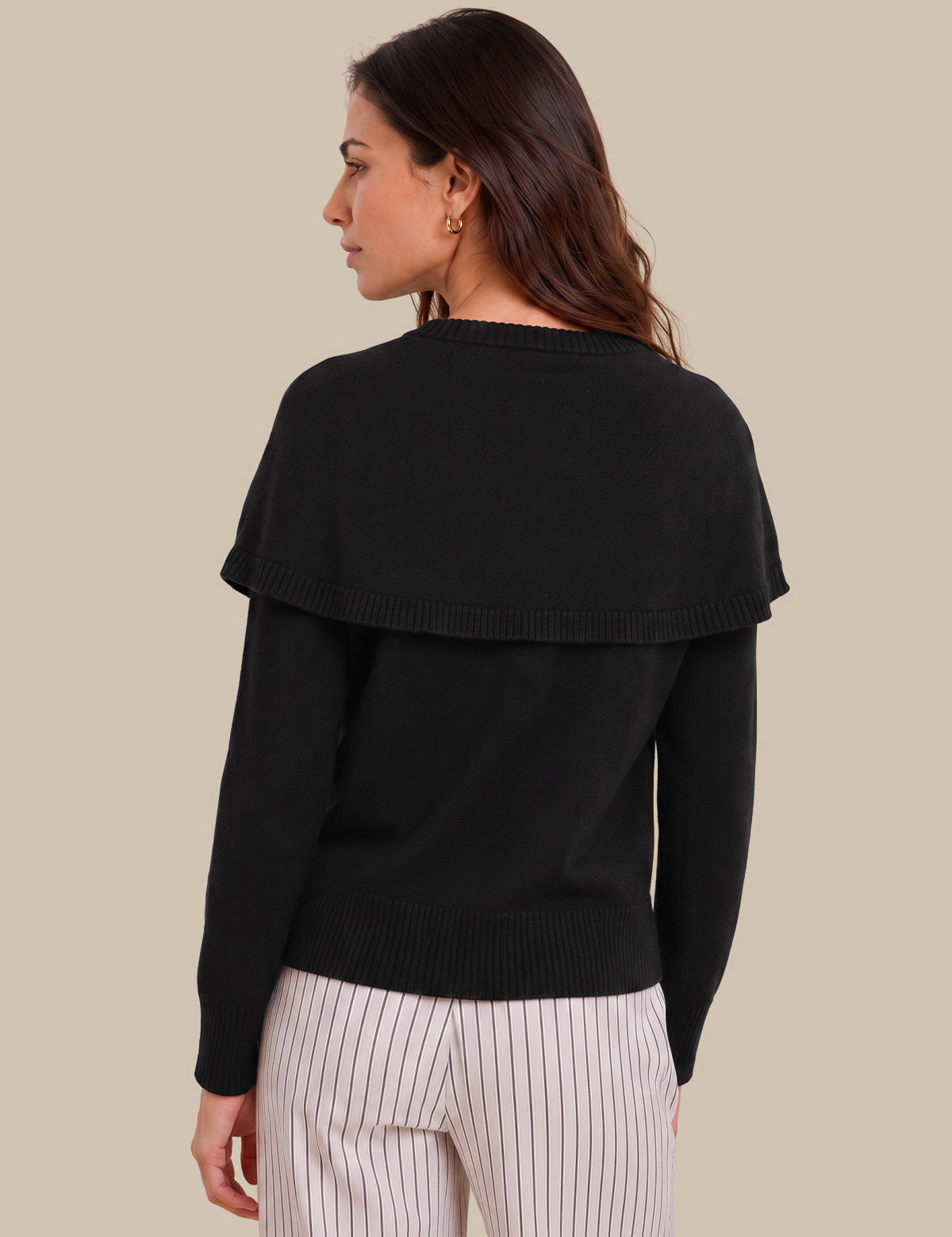 A woman shown from the back wearing a black long-sleeve knit sweater with a wide draped shoulder detail, paired with white pinstriped pants, against a solid beige background.