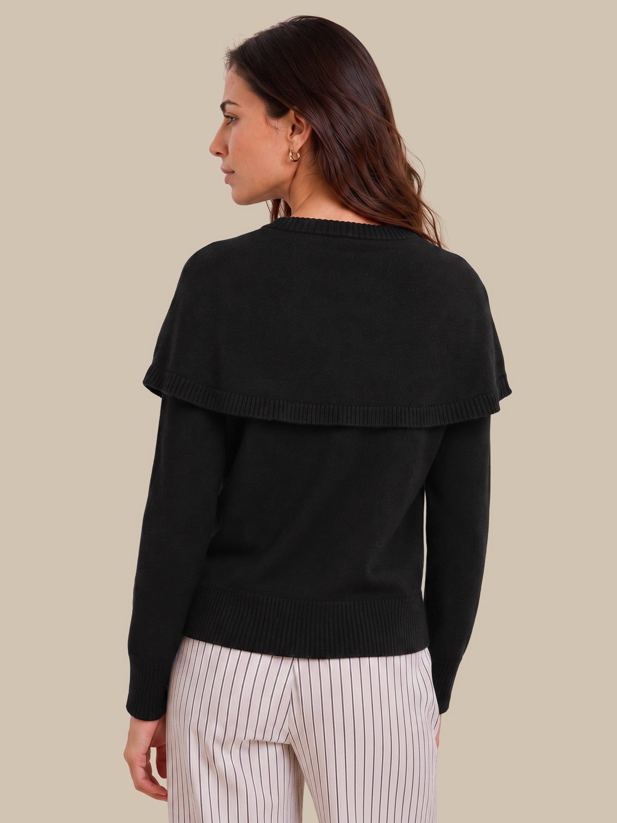 A woman shown from the back wearing a black long-sleeve knit sweater with a wide draped shoulder detail, paired with white pinstriped pants, against a solid beige background.