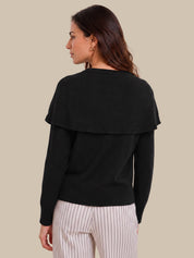 A woman shown from the back wearing a black long-sleeve knit sweater with a wide draped shoulder detail, paired with white pinstriped pants, against a solid beige background.