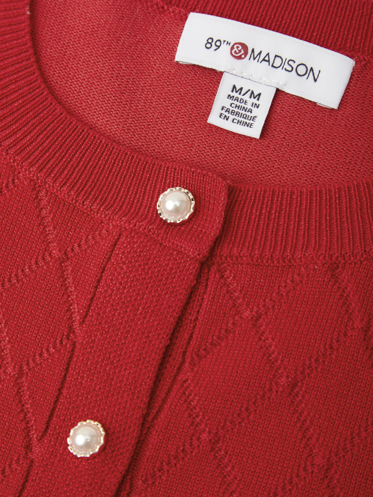 Close-up of a red knit cardigan sweater showing the ribbed collar, textured knit pattern, and two pearl-like buttons. The inner neck label reads 89th & Madison.