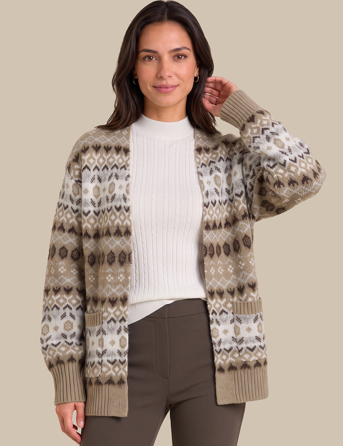 A woman modeling a beige and brown Fair Isle patterned cardigan sweater over a white ribbed top and dark brown pants, set against a solid beige studio background.