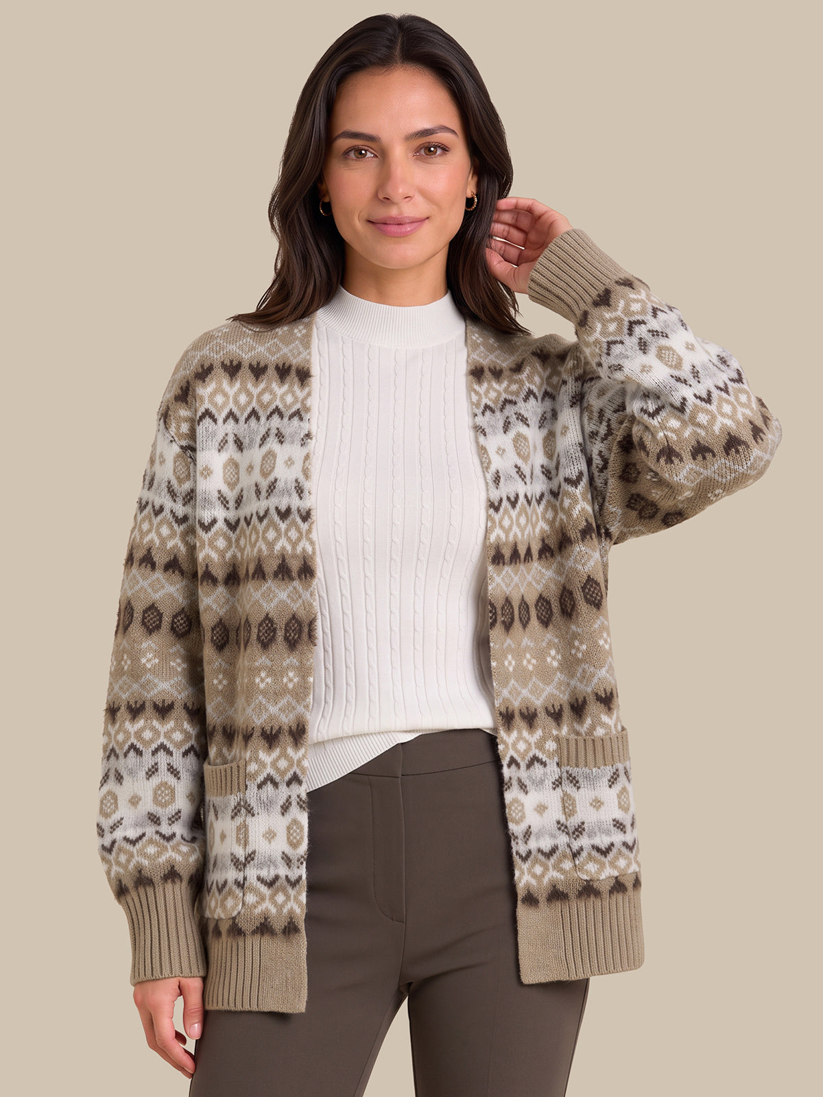 A woman modeling a beige and brown Fair Isle patterned cardigan sweater over a white ribbed top and dark brown pants, set against a solid beige studio background.