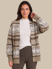 A woman modeling a beige and brown Fair Isle patterned cardigan sweater over a white ribbed top and dark brown pants, set against a solid beige studio background.