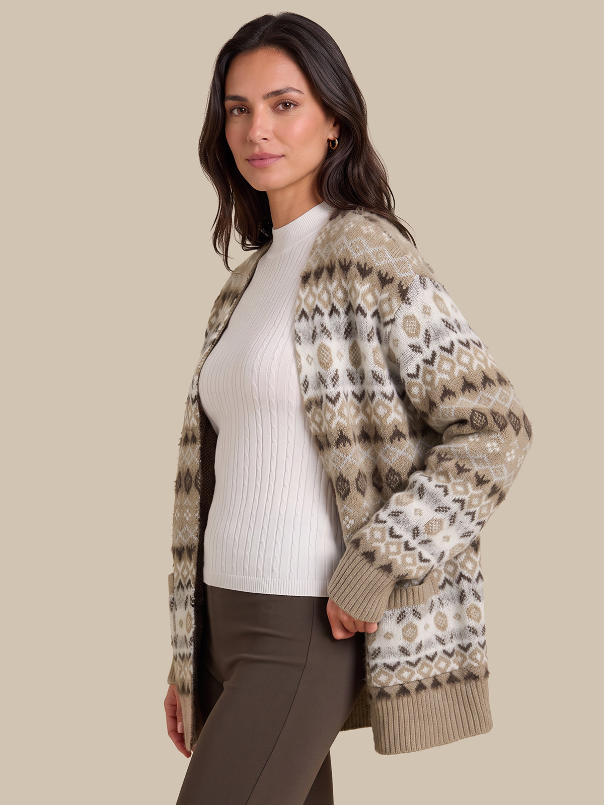 A woman wearing a beige and brown patterned knit cardigan over a white ribbed top and brown pants, photographed against a solid beige studio background.