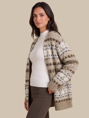 A woman wearing a beige and brown patterned knit cardigan over a white ribbed top and brown pants, photographed against a solid beige studio background.