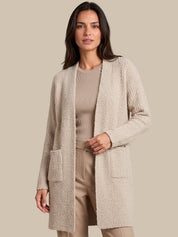 A woman modeling a textured, open-front cardigan in a light beige color, paired with a matching ribbed top and beige trousers, set against a solid beige background.