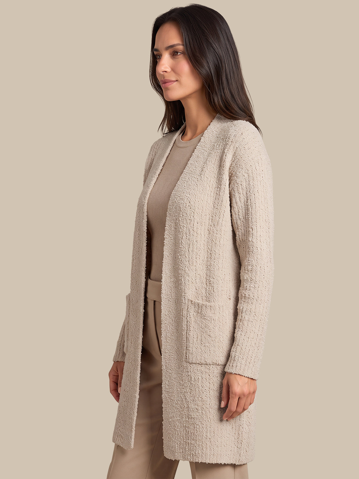 A woman wearing a textured, open-front cardigan in a light beige/taupe color over a matching beige top and trousers, set against a solid beige studio background.