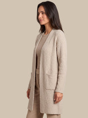 A woman wearing a textured, open-front cardigan in a light beige/taupe color over a matching beige top and trousers, set against a solid beige studio background.
