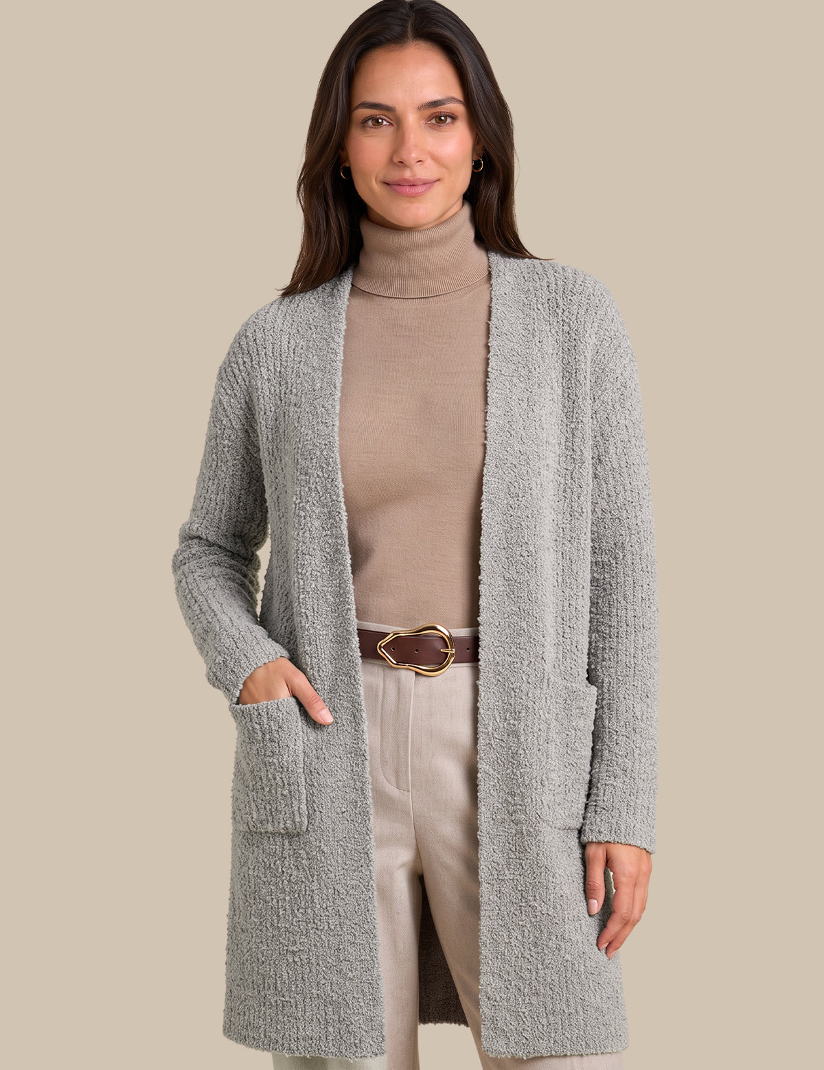 A woman modeling a light gray textured open-front cardigan over a beige turtleneck, cream pants, and a brown belt, set against a solid beige background.