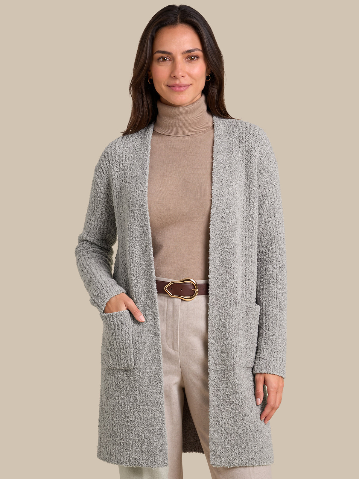 A woman modeling a light gray textured open-front cardigan over a beige turtleneck, cream pants, and a brown belt, set against a solid beige background.