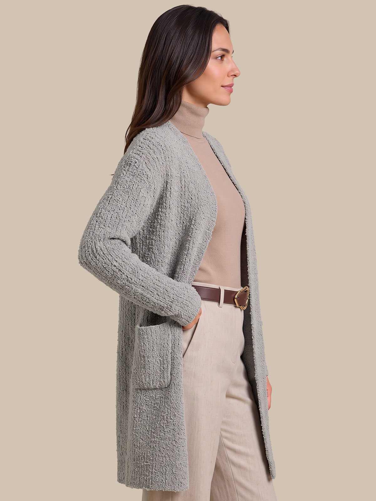 A woman shown in side profile wearing a textured light gray open-front cardigan over a beige turtleneck, paired with beige pants and a brown belt, against a solid beige studio background.