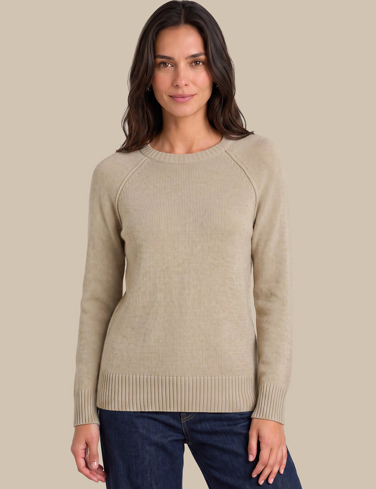A woman wearing a light beige ribbed knit sweater with raglan sleeves and dark blue jeans, standing against a solid beige background.