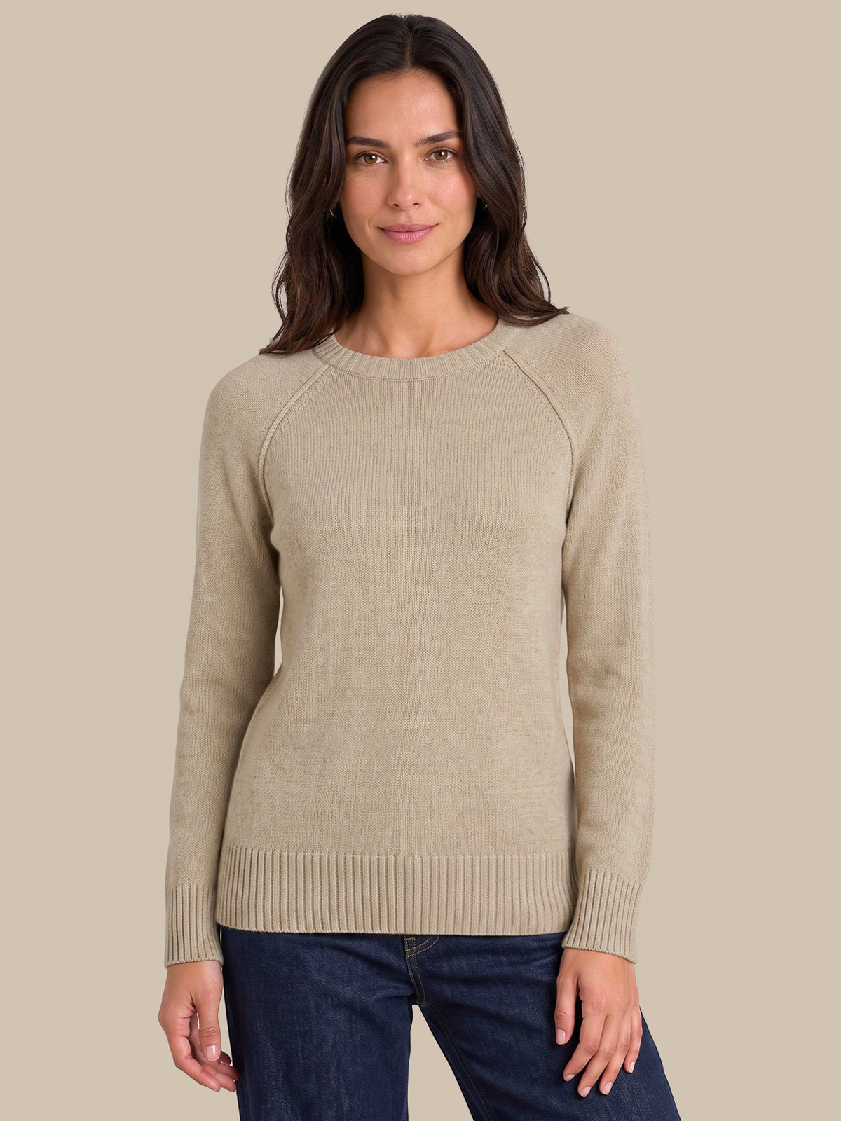 A woman wearing a light beige ribbed knit sweater with raglan sleeves and dark blue jeans, standing against a solid beige background.