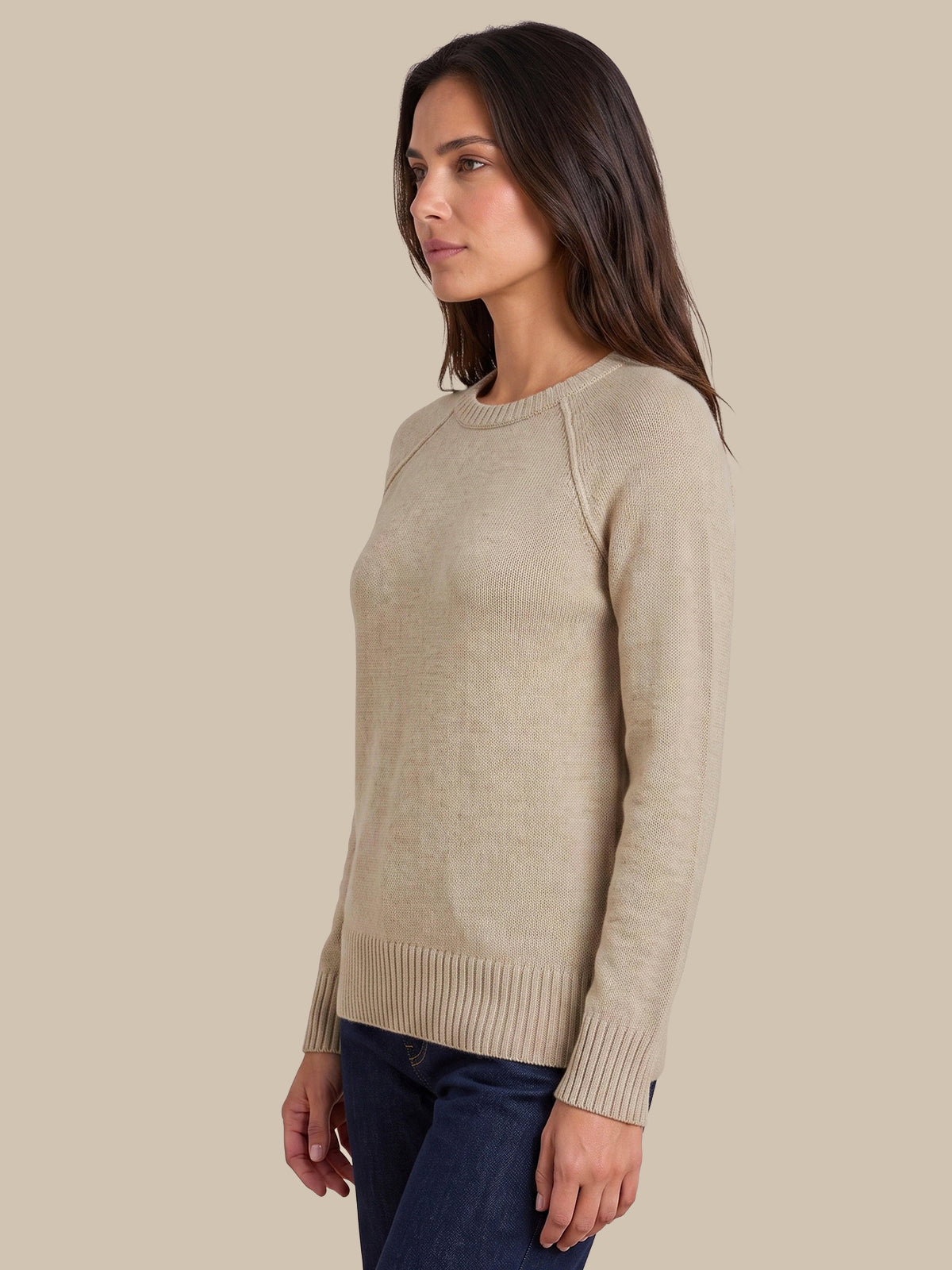 A woman wearing a light beige crewneck knit sweater and dark blue jeans, shown in a side profile against a solid beige background.